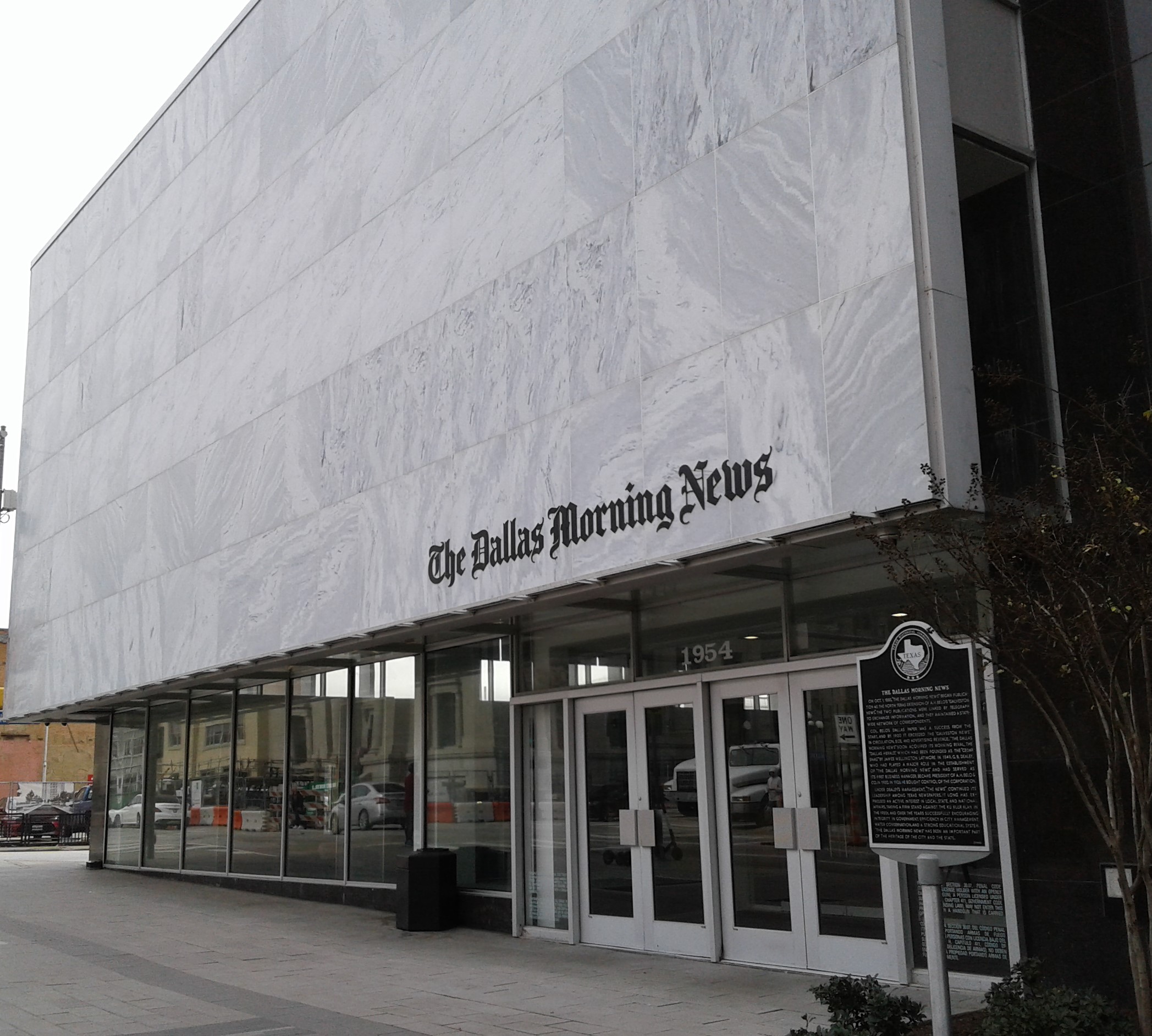 This image shows the facade of 'The Dallas Morning News' building with large windows, a historical marker, and a white marble exterior with signage.