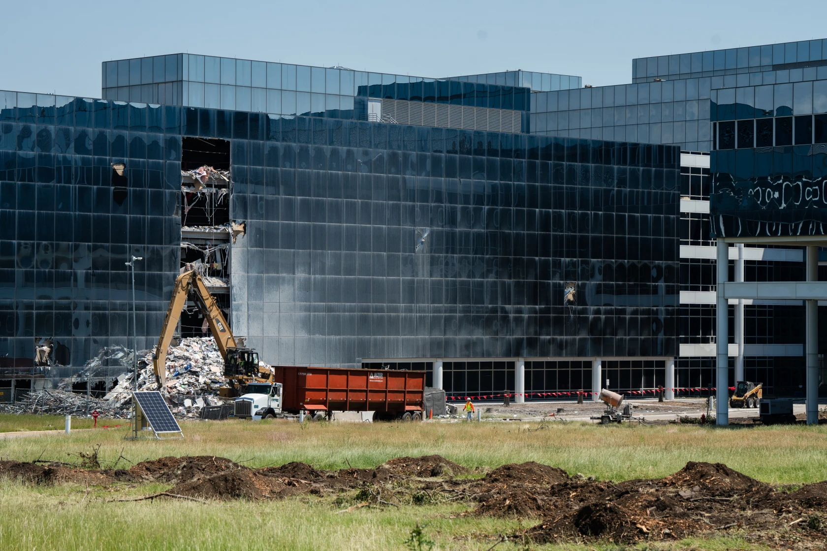 A damaged modern building with exposed interiors, an excavator at work, debris in a dumpster, and a person in safety gear on site.