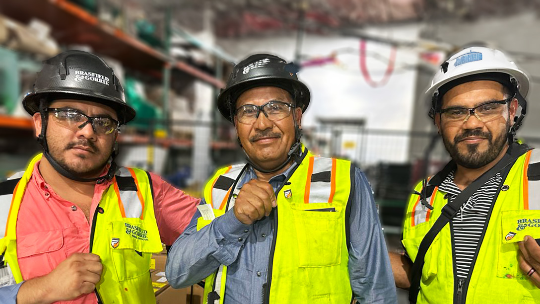 Frontline Leadership Program: Three people in hard hats and high-visibility vests stand confidently in an industrial setting with shelves and equipment blurred in the background.